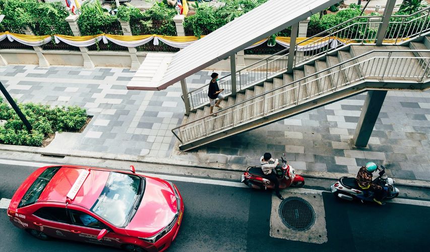 man walks on footbridge with two motorcycles illegally parked under footbridge with red car waiting behind