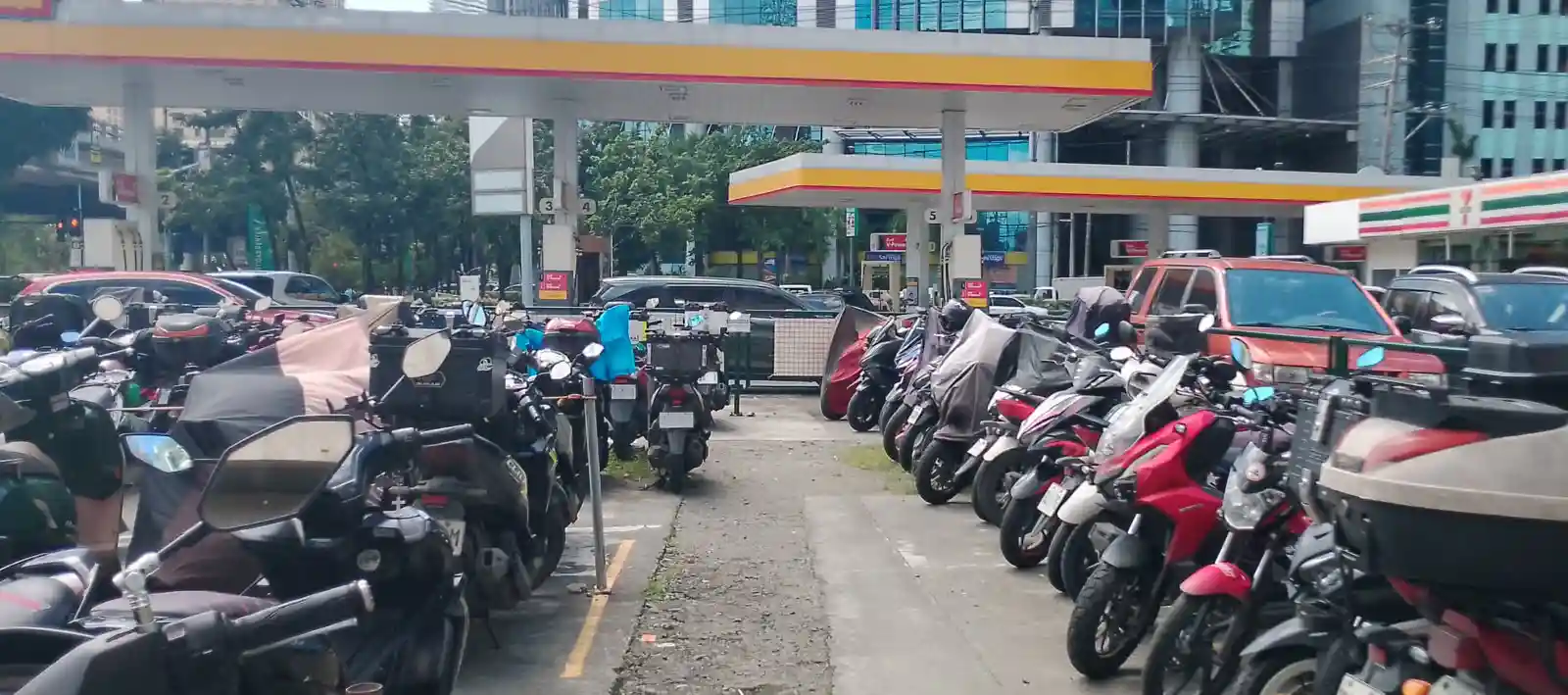 Rows of parked motorcycles fill the foreground near a Shell gas station, with cars and commercial buildings visible in the background on a sunny day.