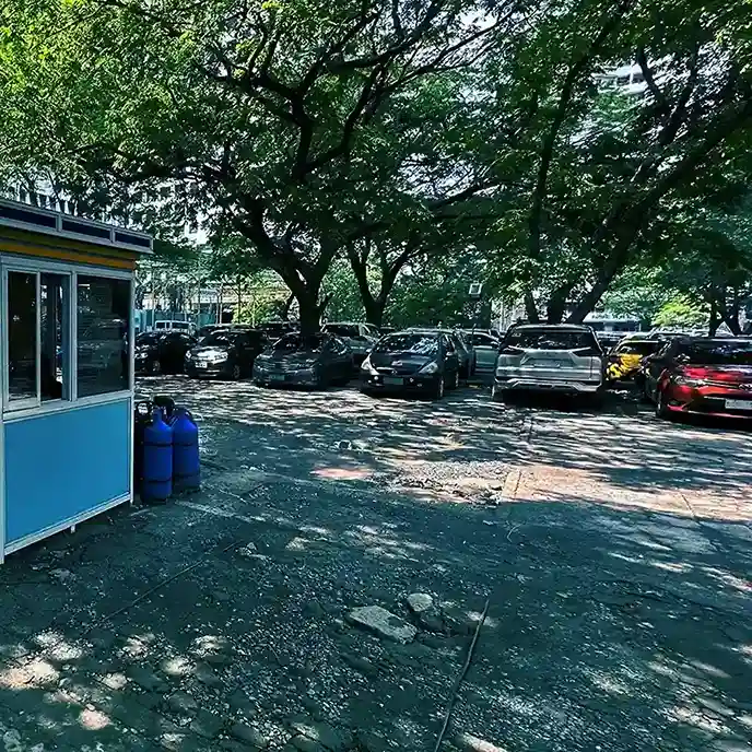 A shaded parking lot with several cars parked under large leafy trees. A small blue and white guard booth stands on the left side of the image, with two blue gas cylinders beside it. The ground is uneven and cracked, with sunlight filtering through the trees.