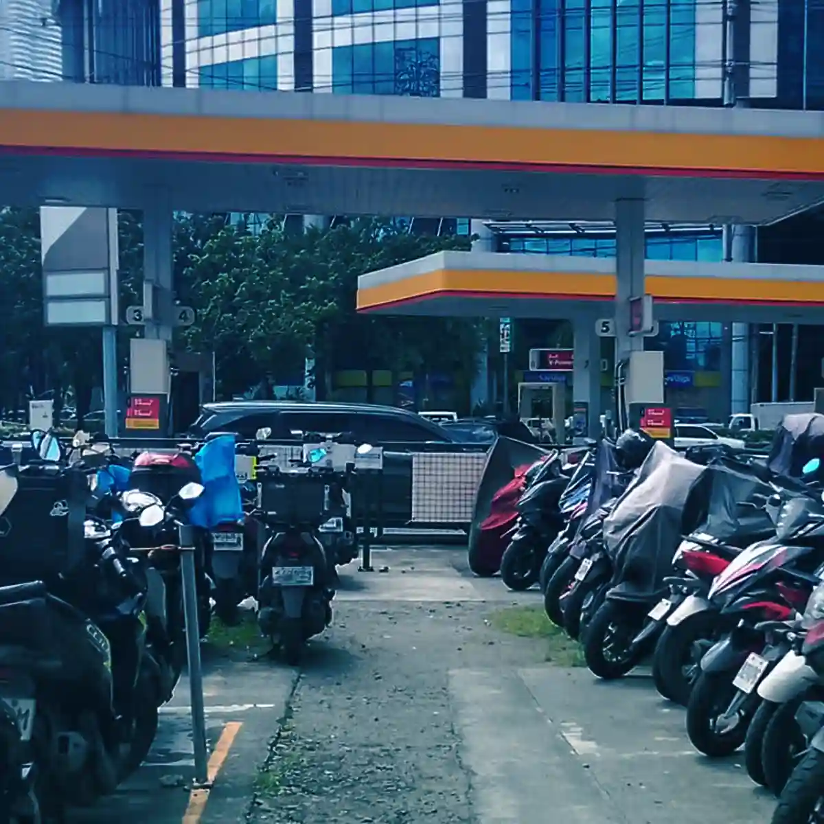 A parking area filled with motorcycles lined up on both sides of a narrow path. In the background, a gas station with orange and yellow signage is visible, along with several cars and a tall glass office building.