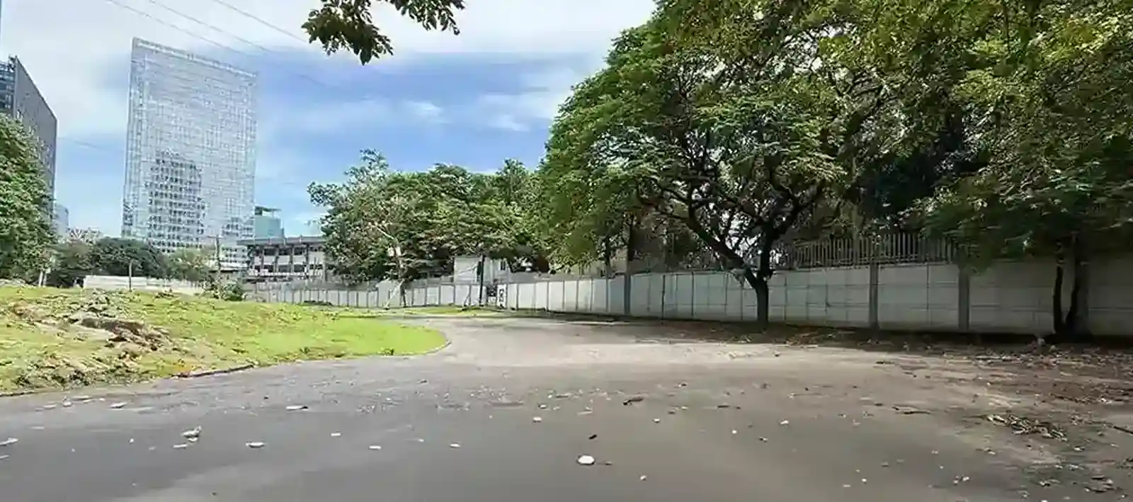 A curved paved pathway bordered by grass and large leafy trees, with tall modern buildings visible in the background under a partly cloudy sky.