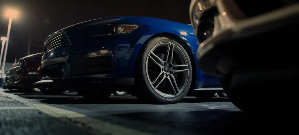 Close-up view of a blue car parked in a lot at night under streetlights, with other vehicles lined up in the background.