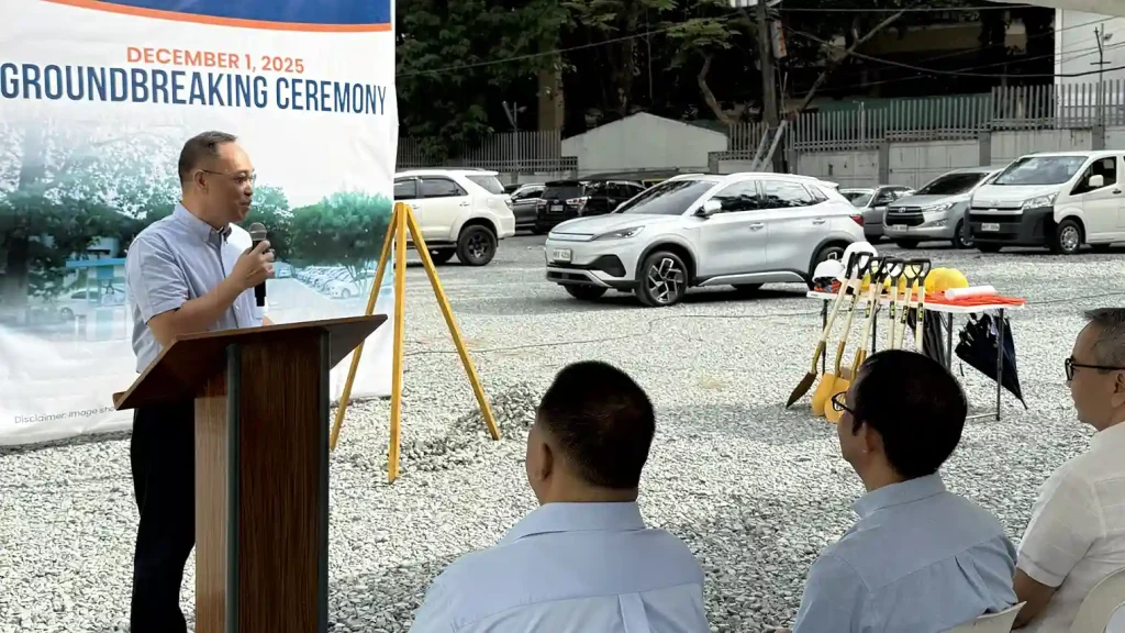 Man speaking at a podium during the December 1, 2025 groundbreaking ceremony, with attendees seated in front, cars parked in the background, and a table holding shovels and safety gear at the construction site